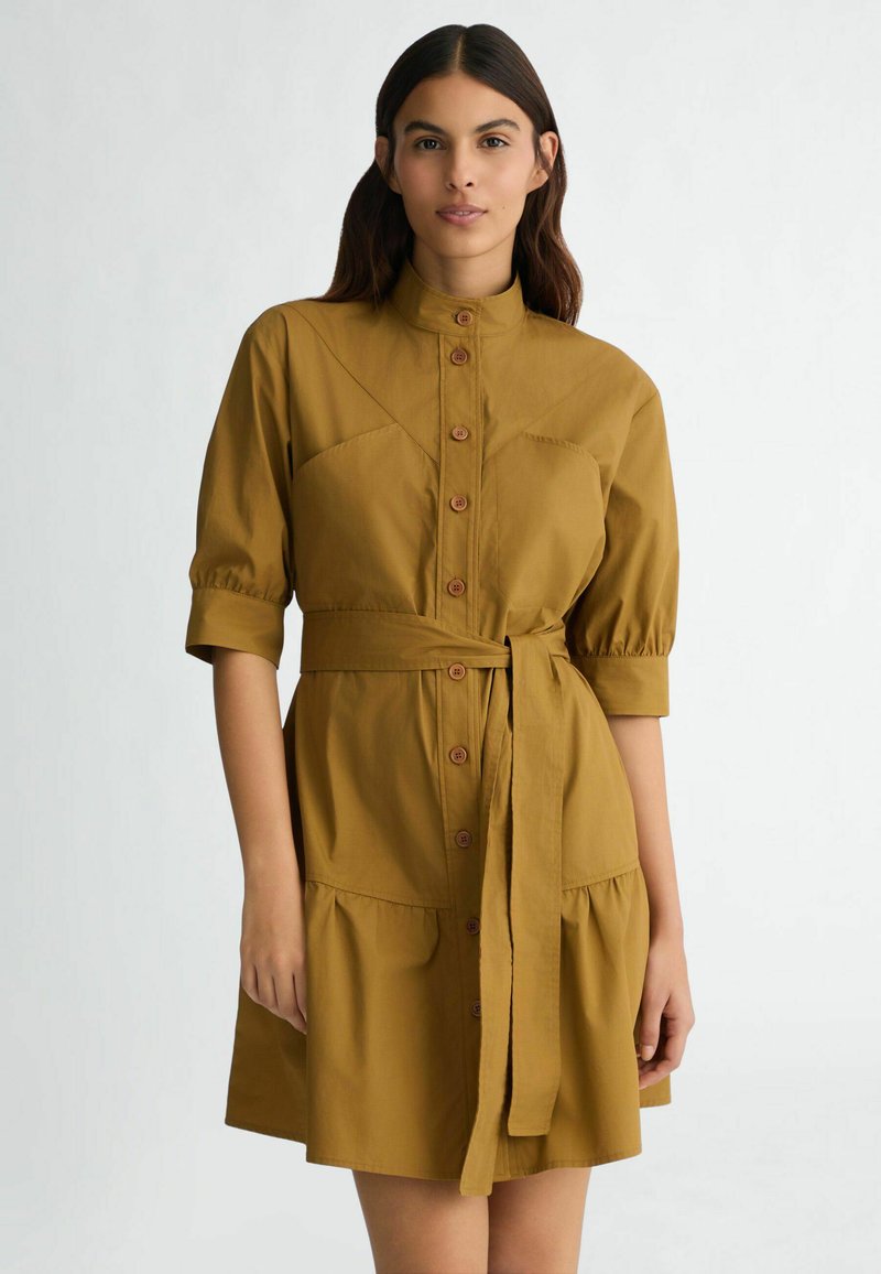 Robe chemise couleur moutarde avec col, fermeture à boutons sur le devant, manches bouffantes, taille ceinturée et jupe à volants. Texture de tissu lisse.