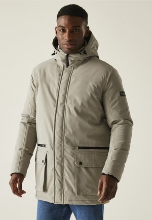 Chaqueta acolchada beige con capucha, que cuenta con una cremallera frontal, dos bolsillos laterales con cremallera y un bolsillo con parche. Textura suave con acentos en negro.