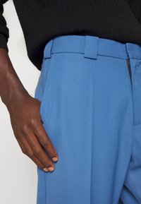 Persona dalla pelle scura che indossa pantaloni sartoriali blu e maglione nero lavorato a maglia, con la mano sinistra appoggiata vicino alla tasca del pantalone.