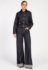 Denimjasje met zwart corduroy kraag, gouden knopen, twee voorkrufen, gecombineerd met denimbroek met wijde pijpen en opgerolde manchetten. Donker gewassen.