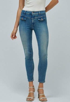 Jeans slim en denim bleu moyen, taille haute avec cinq poches, couture visible et délavage. Associés à des talons beiges à lanières.