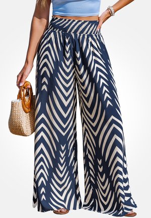 Frau trägt hoch taillierte, weit geschnittene Hosen mit Marine- und Weißem Chevron-Muster, hellblaues Crop-Top, hält gewebte Handtasche, Sandalen sichtbar.