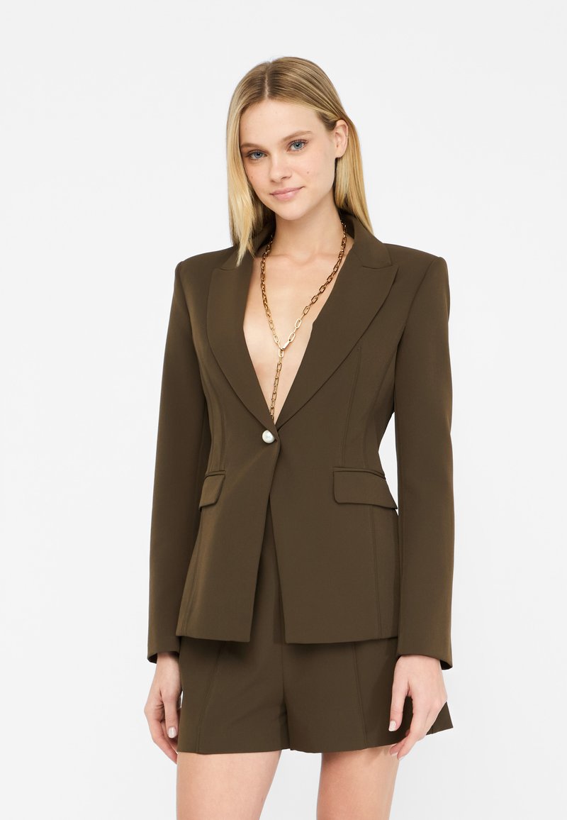 Blazer marrone su misura con scollatura a V profonda, chiusura a un bottone, tasche frontali e silhouette aderente, abbinato a pantaloni corti coordinati.