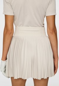 Person som bär en beige plisserad kjol och en ljusbeige kortärmad topp, med en vit handske på vänster hand, visad bakifrån.