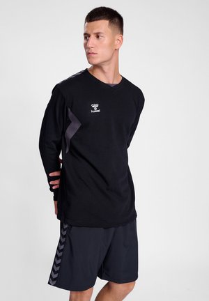 Młody mężczyzna w czarnej sportowej koszulce z długim rękawem i czarnych szortach z wzorzystymi paskami po bokach, patrzy w lewo na jednolitym tle.