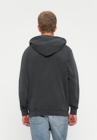 Man with short curly hair wearing a dark gray hoodie and light blue jeans, standing facing a plain white background, back visible.