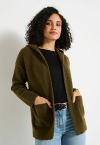 Cardigan en tricot vert olive avec capuche, deux poches avant et manches longues, porté par-dessus un top noir et un jean bleu. Texture douce et coupe décontractée.