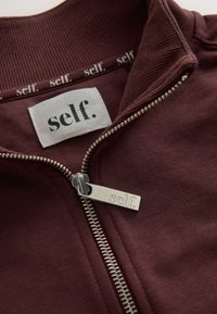 Close-up of a brown zip-up garment with a silver zipper pull and clothing label both engraved with the word "self."