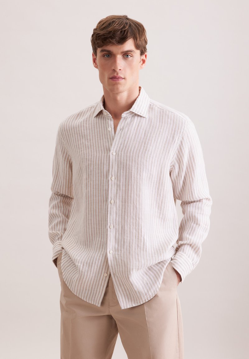 Jeune homme portant une chemise à rayures verticales beige et blanche boutonnée, avec un pantalon beige, debout, les mains dans les poches, sur un fond uni.