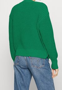 Pull en tricot vert avec un motif texturé, des poignets et un ourlet côtelés, présentant des fentes sur les côtés et une coupe décontractée, associé à un jean bleu.