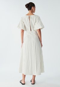 White, knee-length dress featuring black polka dots, flared sleeves, and a keyhole back with zipper. Smooth texture and pleated design.