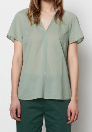 Femme portant un blouse à manches courtes en V vert clair et un pantalon vert foncé, se tenant devant un fond blanc uni.