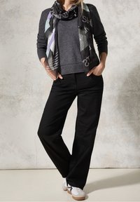 Personne portant un pull gris foncé, un pantalon large noir, des baskets blanches et une écharpe noire à motifs, debout devant un mur à texture neutre.