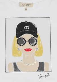 Biała koszulka z grafiką przedstawiającą kobietę z blond włosami w czapce i okrągłych okularach przeciwsłonecznych, ozdobioną kryształkami.