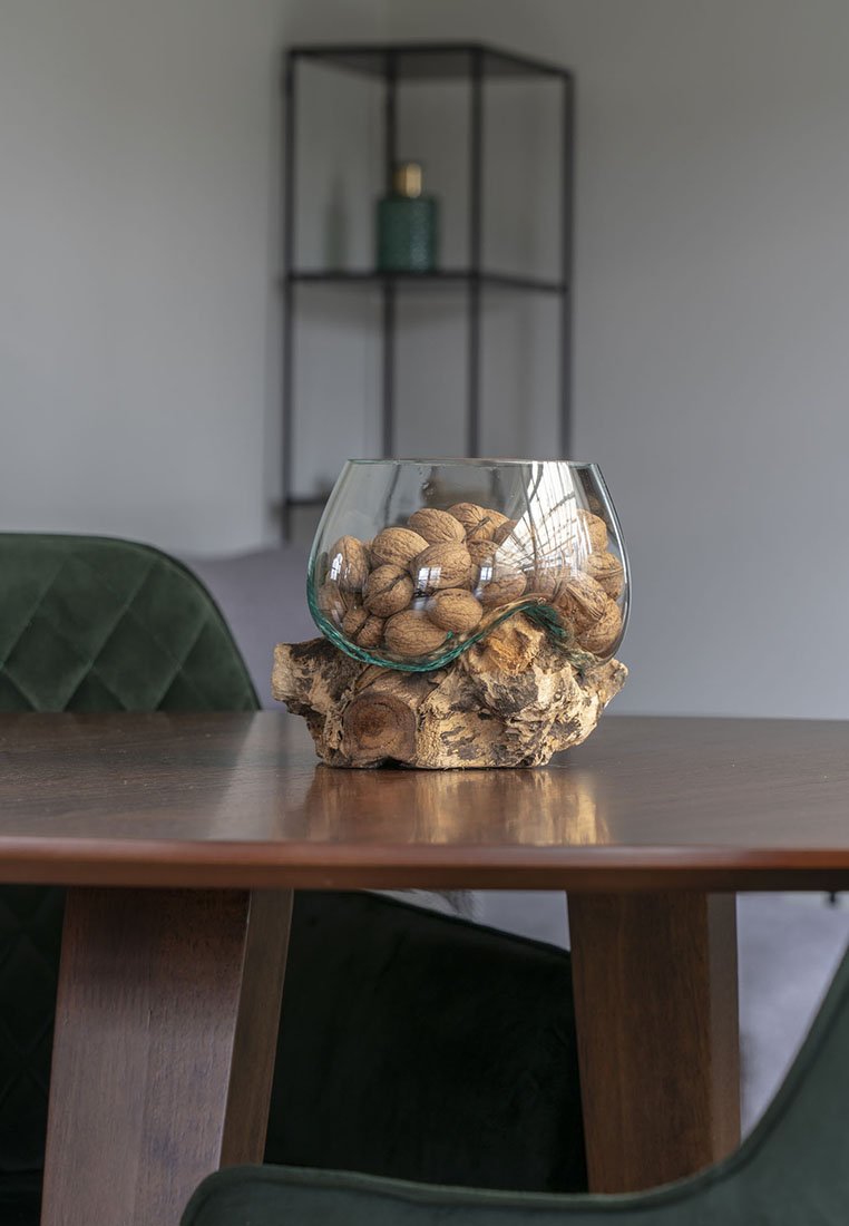 Bol en verre contenant des noix, posé sur une base en bois naturel, sur une table ronde en bois. Chaises rembourrées vertes en arrière-plan, au design minimaliste.