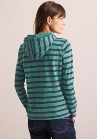 Felpa a righe verde e blu navy con maniche lunghe e cappuccio regolabile. Tessuto morbido, vestibilità comoda, abbinata a jeans scuri.