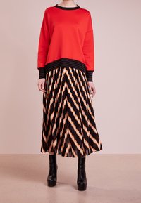Sudadera roja con dobladillo de rayas negras, combinada con una falda plisada con un patrón en chevron en negro, naranja y crema. Botas de plataforma negras.