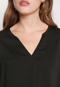 Kvinna som bär guldspiralhängaren och en svart V-ringad topp, med synlig nyckelben och ljusbrunt hår.