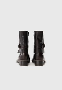 Par de botas de cuero marrón oscuro hasta el tobillo con correas y hebillas, vistas desde atrás sobre un fondo blanco liso.