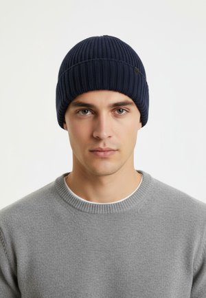 Navy knitted beanie with ribbed texture, featuring a folded cuff and subtle logo accent. Worn with a light gray sweater.