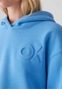 Žena v světlomodrém mikině s vyvýšenými geometrickými tvary na hrudi, včetně kruhu a dvou trojúhelníků, detailní pohled.