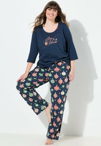 Mujer sonriente con un conjunto de pijama azul marino con el texto "Alegría para el Mundo" en la parte superior y un estampado de adornos navideños coloridos en los pantalones, de pie descalza.