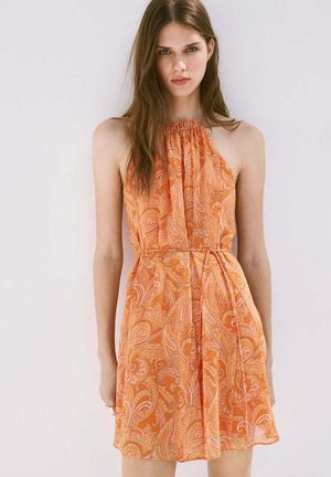 Joven mujer con un vestido naranja sin mangas con estampado cachemira y cuello halter, de pie frente a un fondo blanco liso.