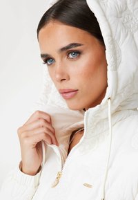 White quilted hooded jacket with embroidered circular patterns, zipped front, gold hardware accents, and drawstring on the hood. Smooth texture.