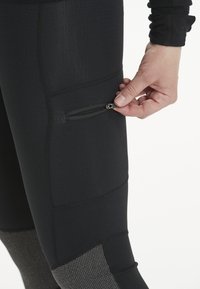 Mustat urheiluhousut, joissa on vetoketjullinen tasku reidessä ja teksturoidut harmaat paneelit alaosassa, valmistettu venyvästä kankaasta.