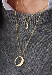 Goldene Halsketten mit einem Halbmond-Anhänger mit einer gehämmerten Textur und einem runden Anhänger, die vor einem grauen Strickpullover präsentiert werden.