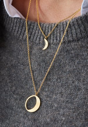 Gouden halskettingen met een halve maan hanger met een gestructureerde afwerking en een ronde hanger, tentoongesteld tegen een grijze gebreide trui.