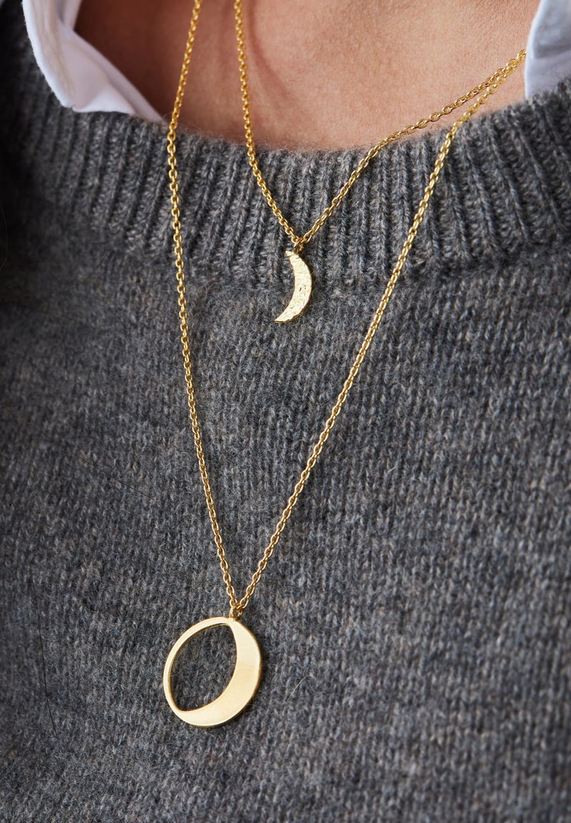 Goldene Halsketten mit einem Halbmond-Anhänger mit einer gehämmerten Textur und einem runden Anhänger, die vor einem grauen Strickpullover präsentiert werden.