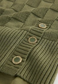 Gros plan sur un tissu tricoté vert avec une texture côtelée et des motifs carrés, comportant trois boutons décoratifs verts et dorés.