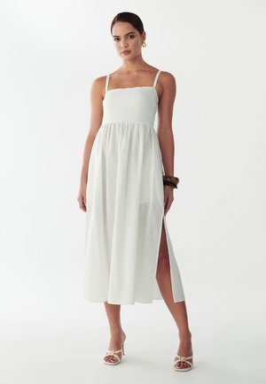 Kvinde iført hvid ærmeløs midikjole med sideslids, beige højhælede sandaler, guldringe og brune armbånd, stående mod hvid baggrund.