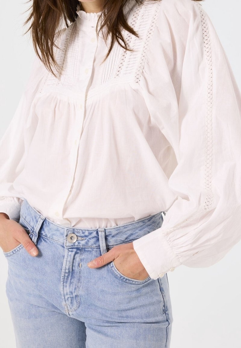 Femme portant un chemisier blanc à manches longues avec des détails en dentelle et un jean bleu clair taille haute, mains dans les poches avant.