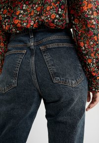 Jeans in denim blu scuro con cinque tasche, cuciture visibili e un taglio dritto. I jeans sono abbinati a una blusa con motivo floreale.