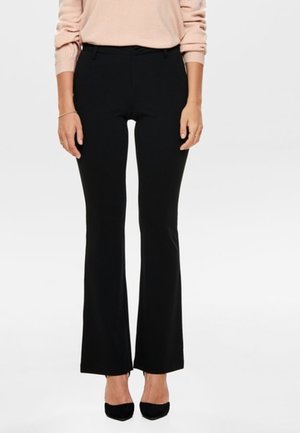 Kobieta ubrana w dopasowane czarne rozszerzane spodnie, zestawione z czarnymi butami na obcasie o spiczastym nosku oraz bladoróżową, długorękawową bluzkę, stojąca na jednolitym tle.