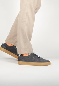 Zapatillas de ante gris oscuro con suela de goma, detalles en blanco y cordones redondos, combinadas con pantalones rectos de color beige claro.