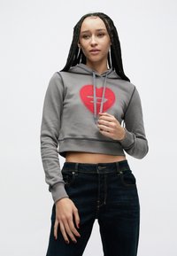 Šedá skrátená mikina s kapucňou so vzorom červeného srdca zdobená trblietkami. Nosená s tmavými džínsami.