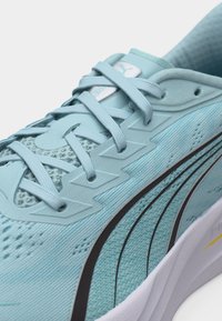 Hellblauer Sportschuh mit Mesh-Obermaterial, Zickzack-Schnürsenkelschlaufen, schwarzem, geschwungenem Streifendesign und weißer Sohle mit gelbem Detail.