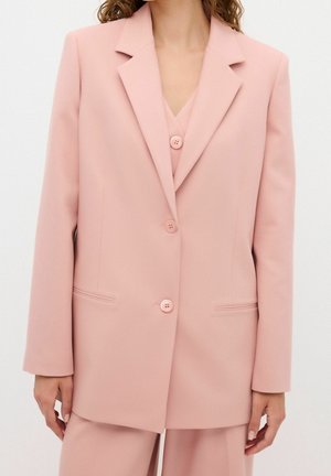 Nainen, jolla on vaaleanpunainen räätälöity bleiseri ja siihen sopivat housut, pitkät hihat ja kolme nappia edessä, seisomassa yksiväristä taustaa vasten.