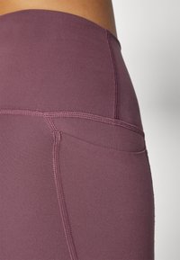 Magas derekú leggings mély lila színben, sima anyaggal, lapos varrással és egy kis oldalsó zsebből.