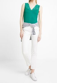 Top sin mangas verde con escote de encaje, pantalones ajustados blancos y una camiseta a rayas atada a la cintura. Combinado con zapatillas blancas.