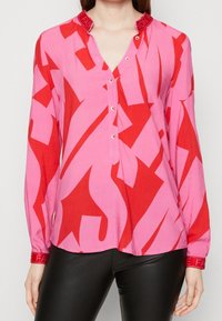 Blusa rosa con un audace motivo astratto rosso, patta con bottoni e accenti di testo rosso a contrasto sui polsini. Tessuto di superficie liscia.