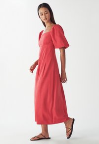 Rdeča maxi obleka z napihnjenimi kratkimi rokavi, kvadratnim izrezom in gubano krilo. Model nosi črne japanke, proti nevtralni ozadju.