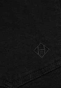 Sort denimstof med en tekstureret overflade; inkluderer et diskret præg af brandlogoet i nederste hjørne. Designet er minimalistisk og strømlinet.