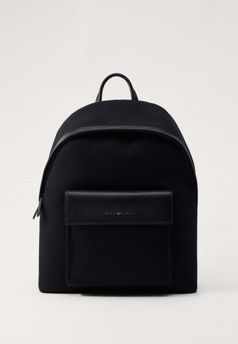Black canvas backpack with a smooth leather front pocket, top handle, and a zipper closure. Features branding on the leather accent.