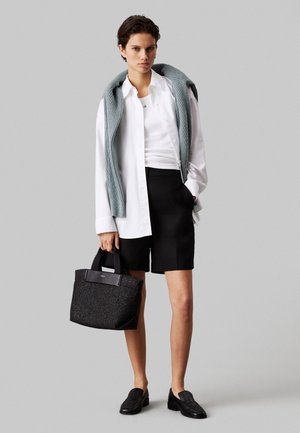 Młoda osoba ubrana w białą koszulę i czarne szorty, szary sweter przewieszony przez ramiona, trzyma czarną torebkę i nosi czarne mokasyny.