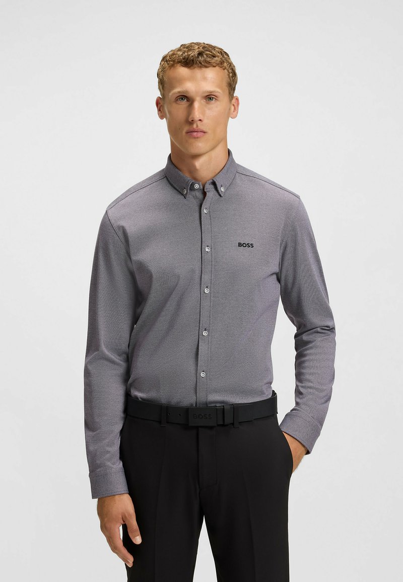 Camicia button-up in tessuto grigio a fantasia con collo, maniche lunghe e un logo discreto sul petto. Abbinata a pantaloni neri.