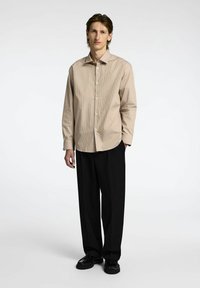Camicia a righe beige con bottoni frontali, maniche lunghe e vestibilità rilassata, abbinata a pantaloni neri a gamba larga e scarpe nere.